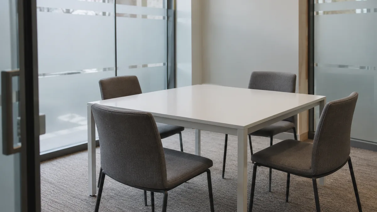 An empty square table and four chairs in a small meeting room with frosted glass walls